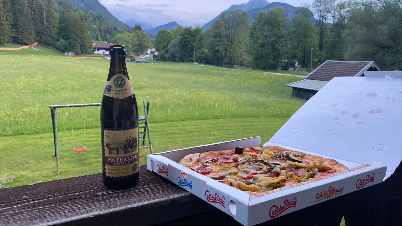Butelka piwa i pizza na balustradzie balkonu, w tle łąka i góry